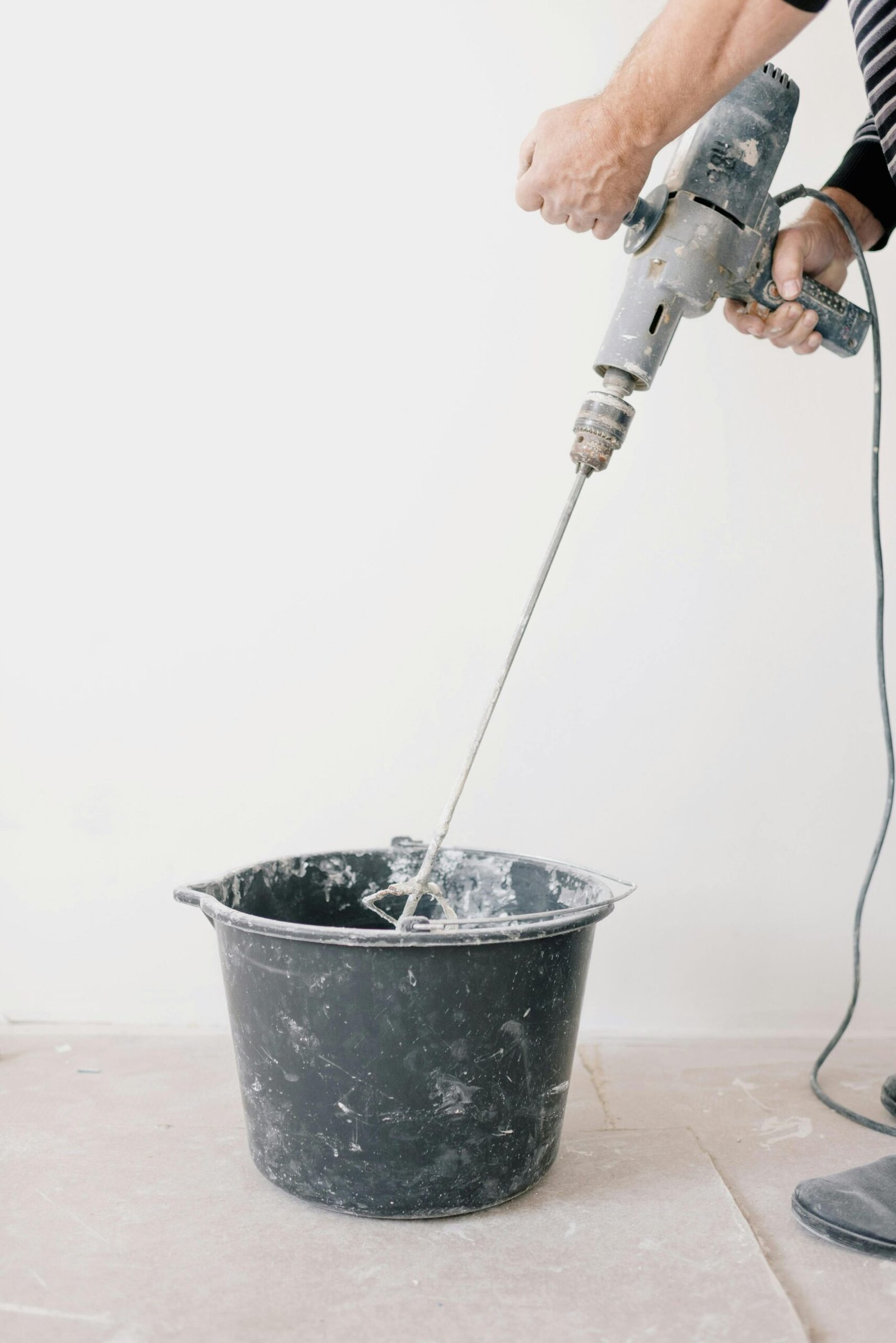 Worker using an electric drill to mix cement in a bucket during home renovation.