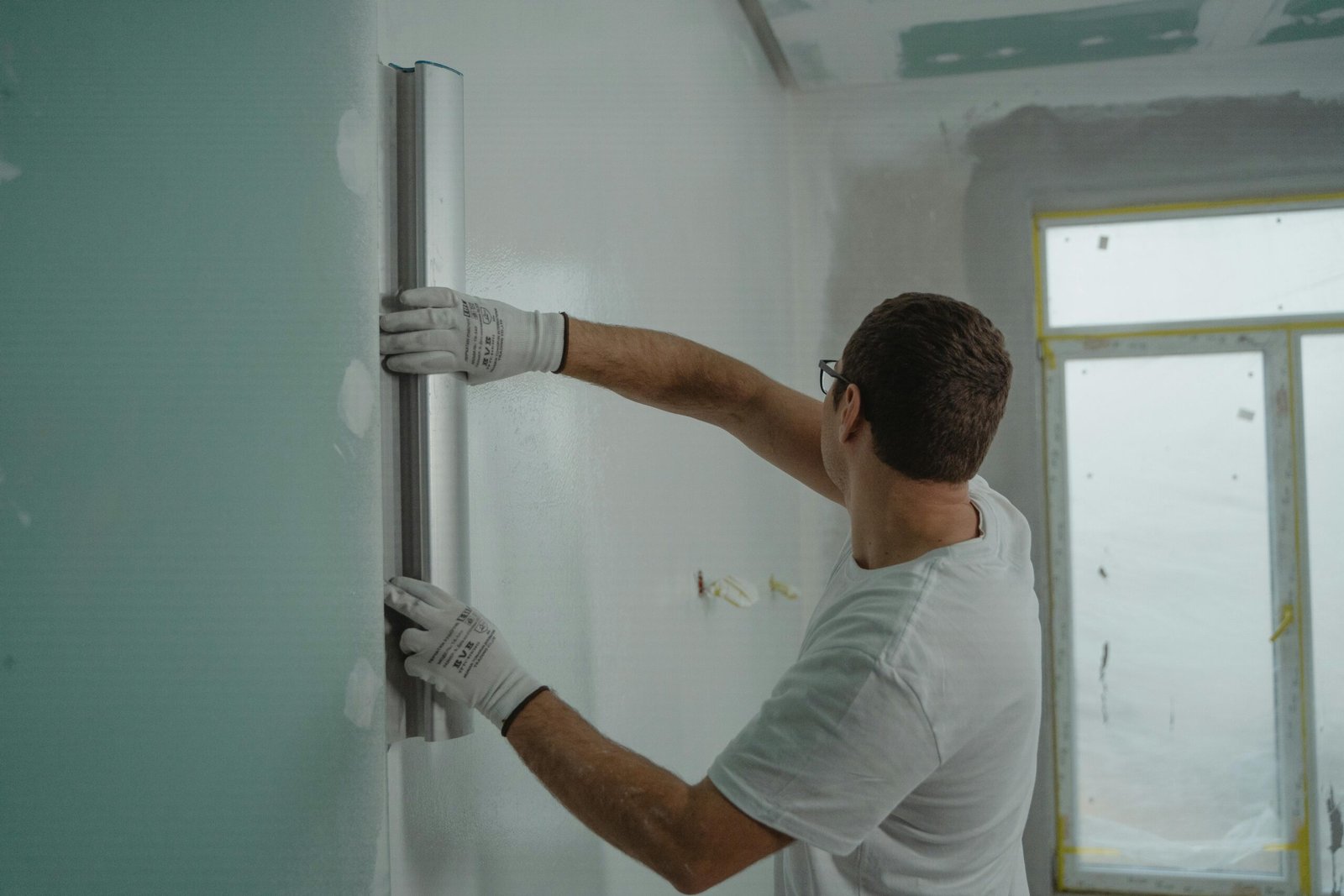 A man in a white shirt and gloves using a metal ruler for indoor wall renovation work.